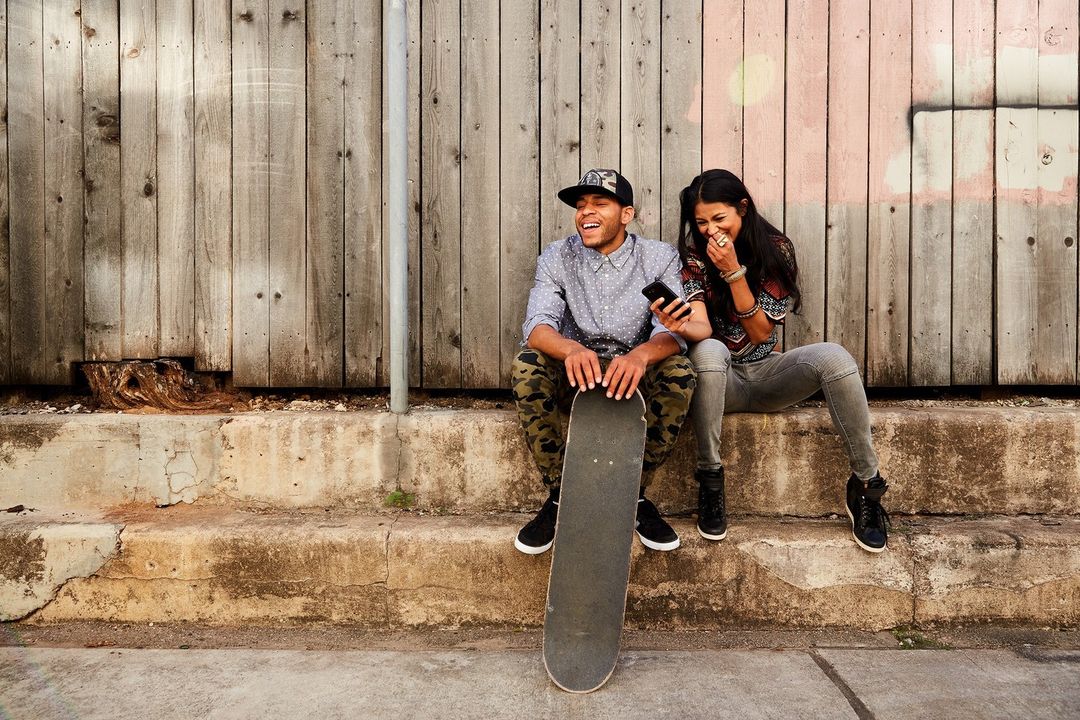 Two friends sitting and laughing near an urban fence with a skateboard. They are dressed casually in daylight.