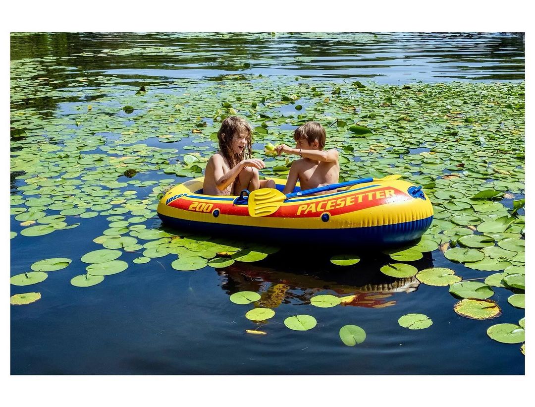 Two kids enjoy a sunny day on the water in an inflatable boat amidst lily pads, with clear water reflecting the boat and children.
