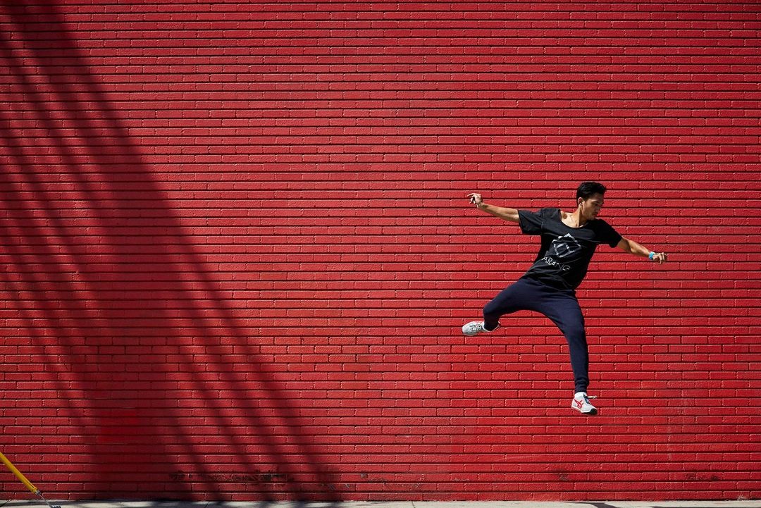 Athletic man in black shirt jumps in the air against a bright red brick wall with shadow patterns on a sunny day.