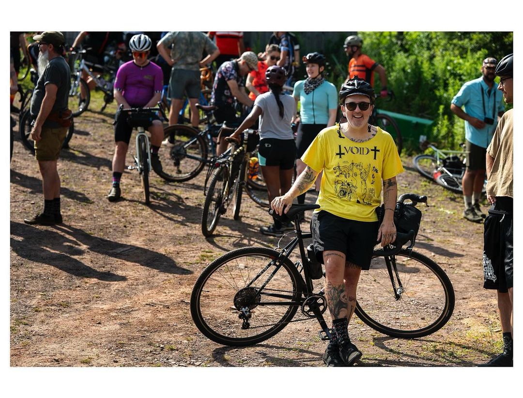 Cyclist in yellow shirt standing with bike among a group of people and bikes on a dirt trail during an outdoor cycling event.