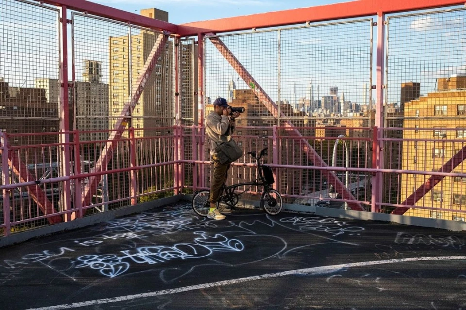 An adult man takes a photograph while standing with his folding bicycle on a pink pedestrian bridge overlooking a city skyline.