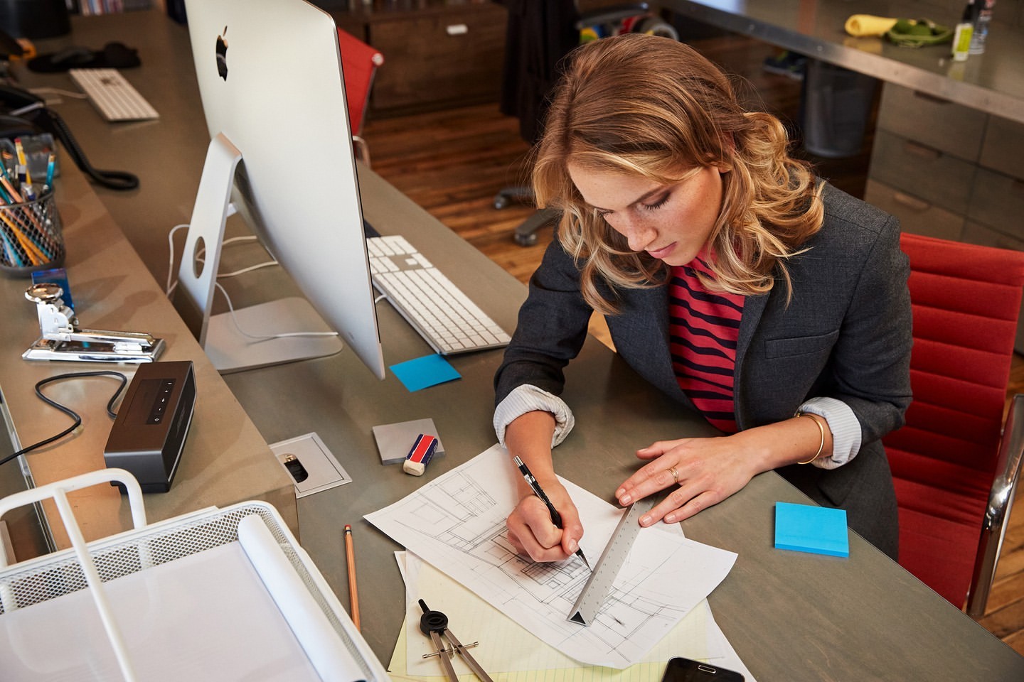 Woman drafting plans at a desk with an Apple iMac computer, using a ruler and pencil with a window view in a modern interior space.