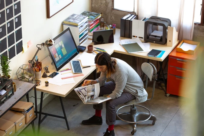 Overhead shot of a person sitting at a desk with a computer, drawing, and office supplies in a modern workspace environment.