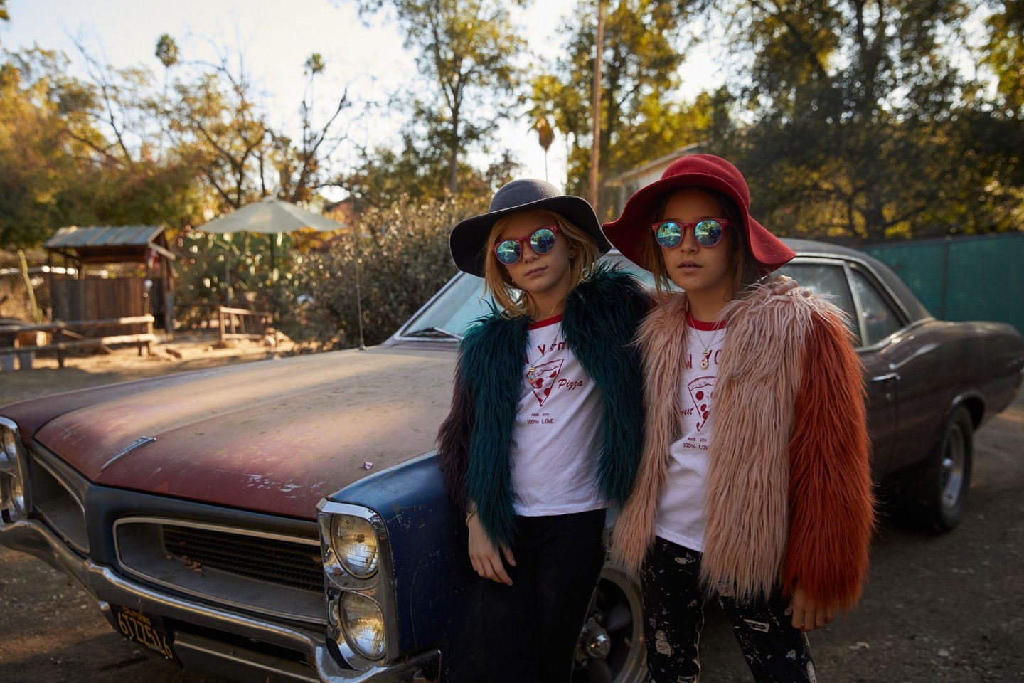 Two young girls in retro sunglasses and hats pose in front of a vintage car on a sunny day in a rural, tree-filled outdoor setting.