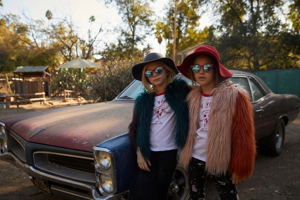 Two young girls in retro sunglasses and hats pose in front of a vintage car on a sunny day in a rural, tree-filled outdoor setting.