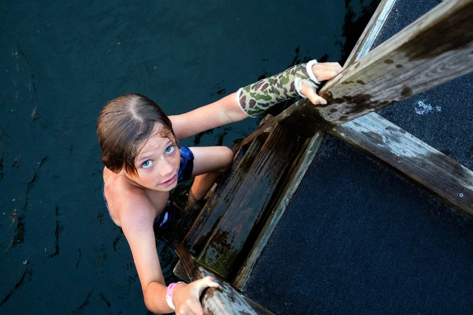 Young boy with a cast climbs a wooden ladder out of dark water, looking upward with wet hair and bright blue eyes in an eye-level shot.