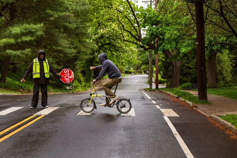 Man rides a small bicycle in a crosswalk as a crossing guard holds up a stop sign in a lush, green suburban neighborhood.