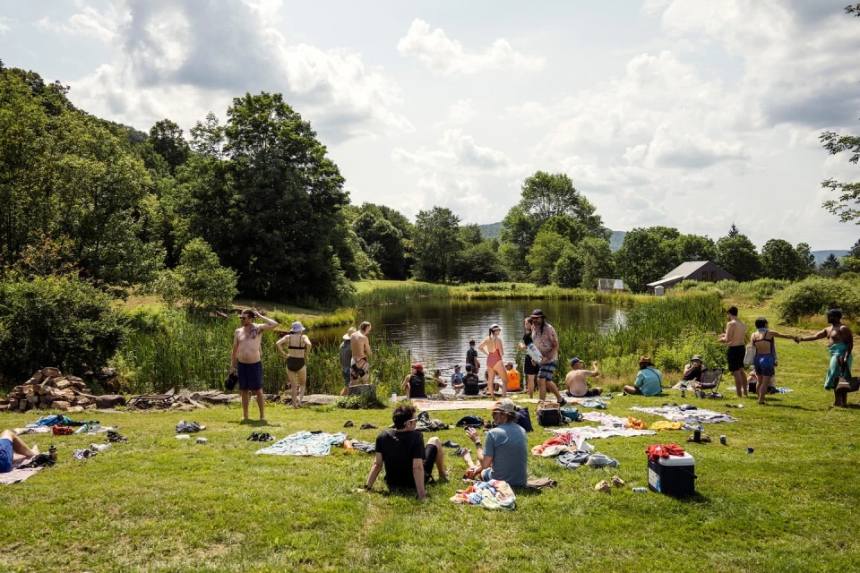 Outdoor gathering on a grassy field next to a pond, with people swimming, sunbathing, and relaxing under a cloudy sky near a forest.