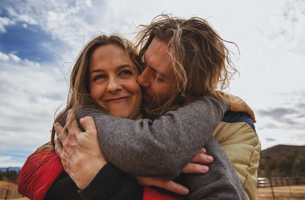 A couple embraces outdoors with a cloudy sky backdrop. An adult male hugs an adult female from behind, kissing her cheek affectionately.