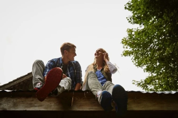 A young couple sitting outdoors on a rooftop ledge with their legs dangling, laughing and looking at each other.