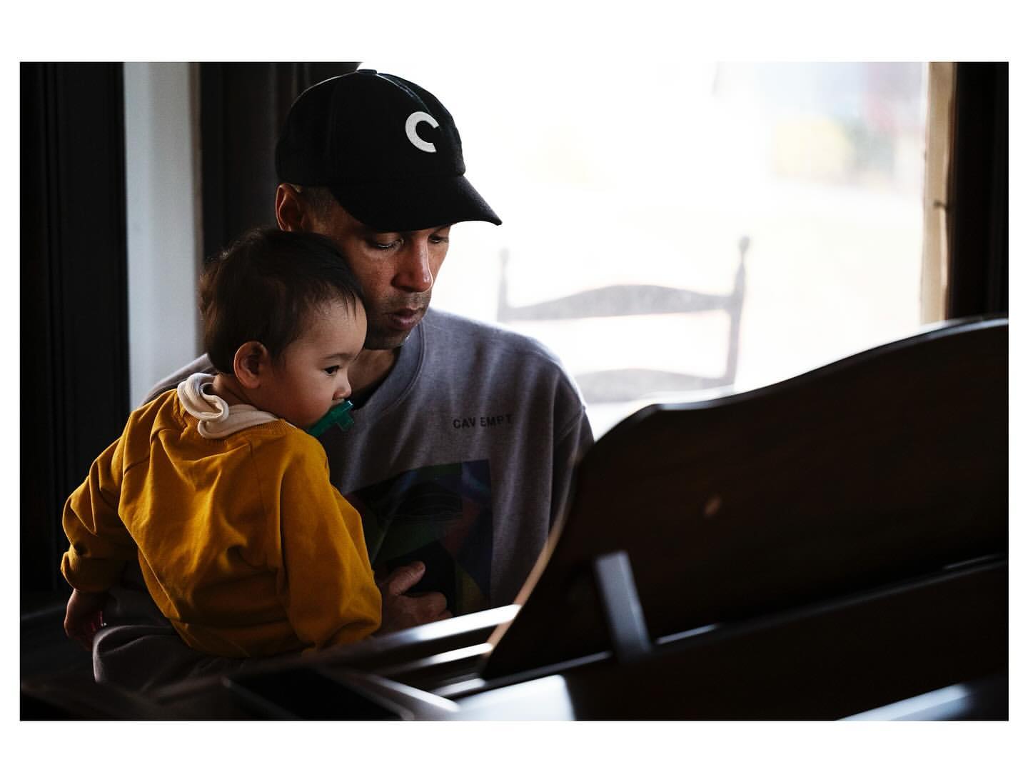A father and child share a quiet moment indoors. The man holds the baby while seated at a piano, both looking towards sheet music.