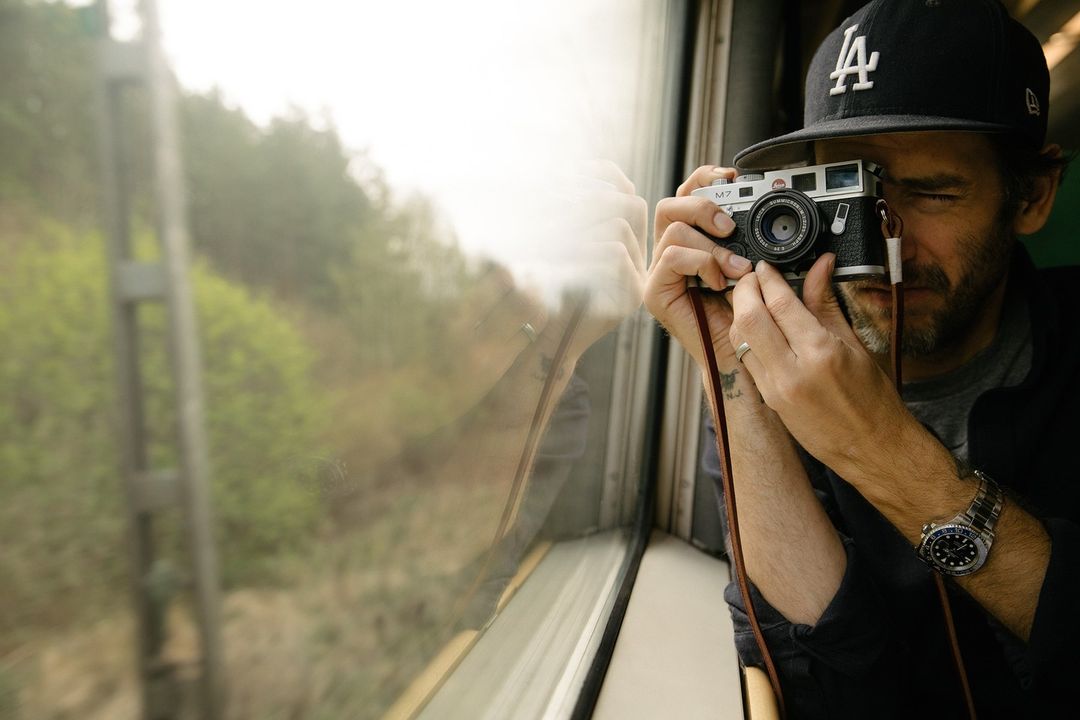 Man wearing an LA hat and wrist watch is sitting inside a train while taking a photo with a Leica M7 camera near window.