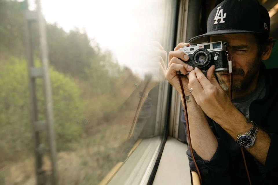 Man wearing an LA hat and wrist watch is sitting inside a train while taking a photo with a Leica M7 camera near window.