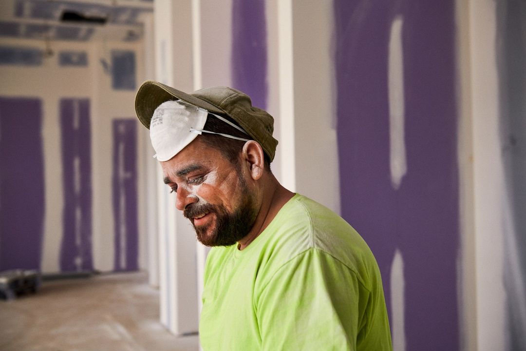 A man in a green shirt and cap, covered in white dust, stands in an unfinished room with unpainted drywall, wearing a face mask.