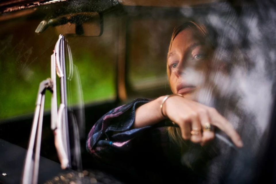 Blonde woman behind the wheel of a car with plaid shirt and jewelry is reflected in the windshield during daytime with green scenery.