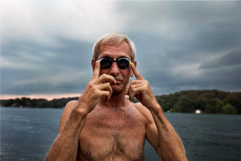 Older man with gray hair and a mustache wearing swimming goggles outdoors with water and trees in the background.