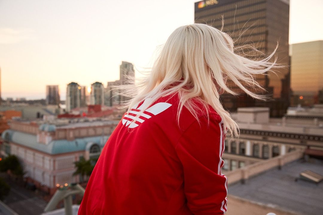Blonde woman in a red Adidas jacket gazes at a city skyline during sunset, creating a vibrant and serene urban scene.