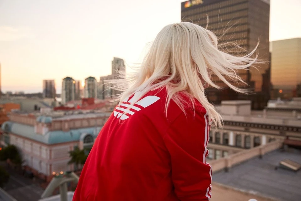 Blonde woman in a red Adidas jacket gazes at a city skyline during sunset, creating a vibrant and serene urban scene.