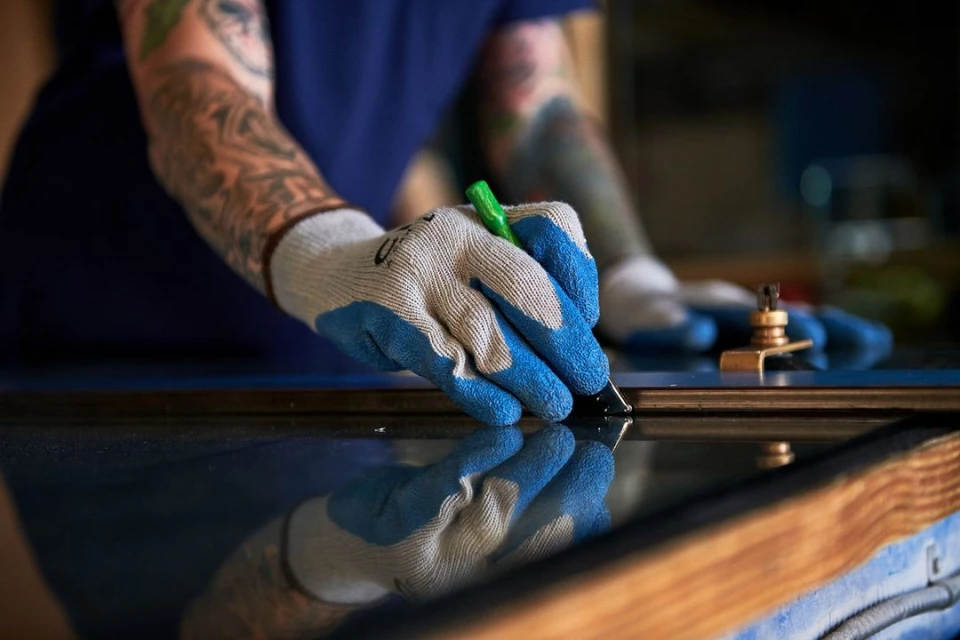 Close-up of tattooed person's gloved hand scoring reflective material with a green-handled tool, craftsmanship visible.