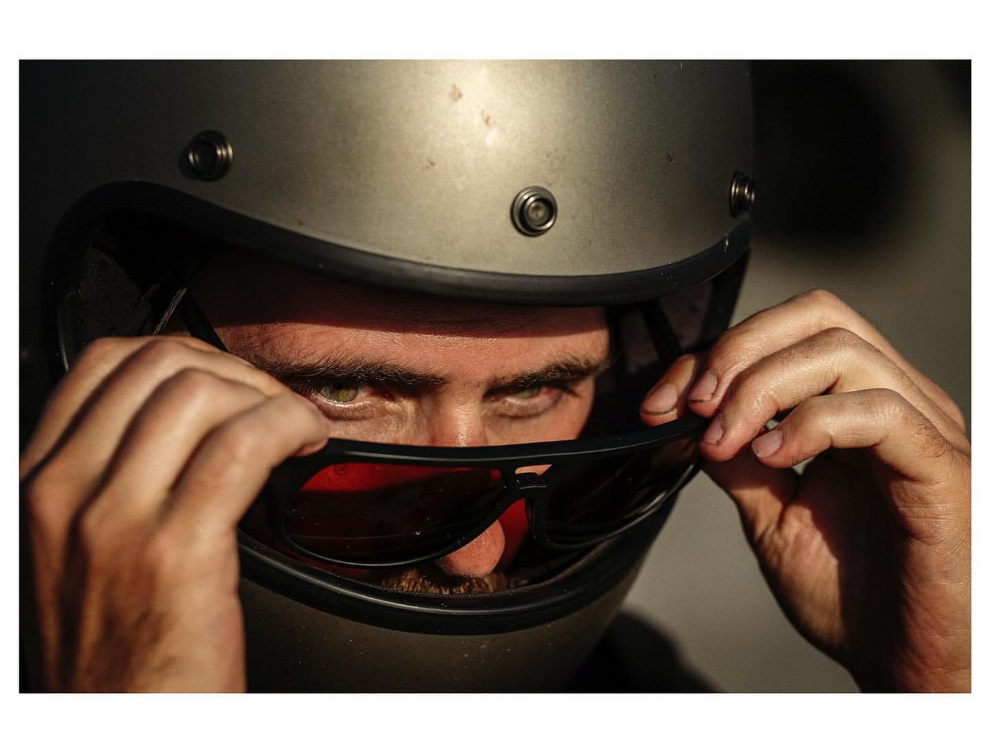 Close-up of an adult male wearing a silver motorcycle helmet and tinted sunglasses, adjusting the sunglasses with both hands.