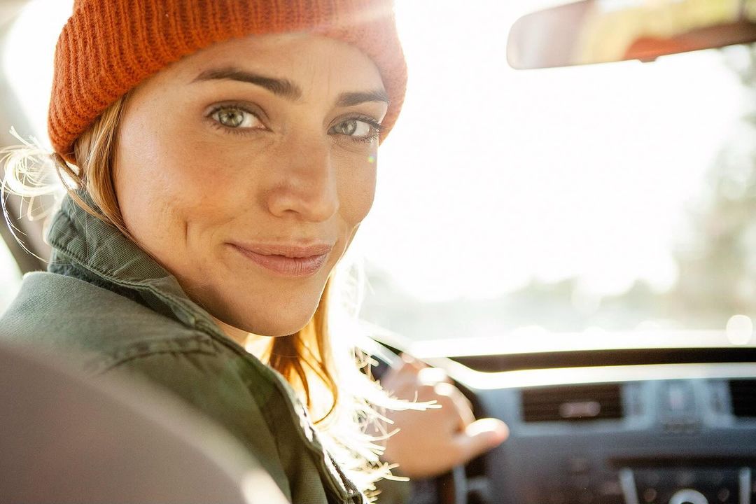 Portrait of a young woman with a beanie, driving and looking at the camera inside a car, bathed in bright natural lighting.