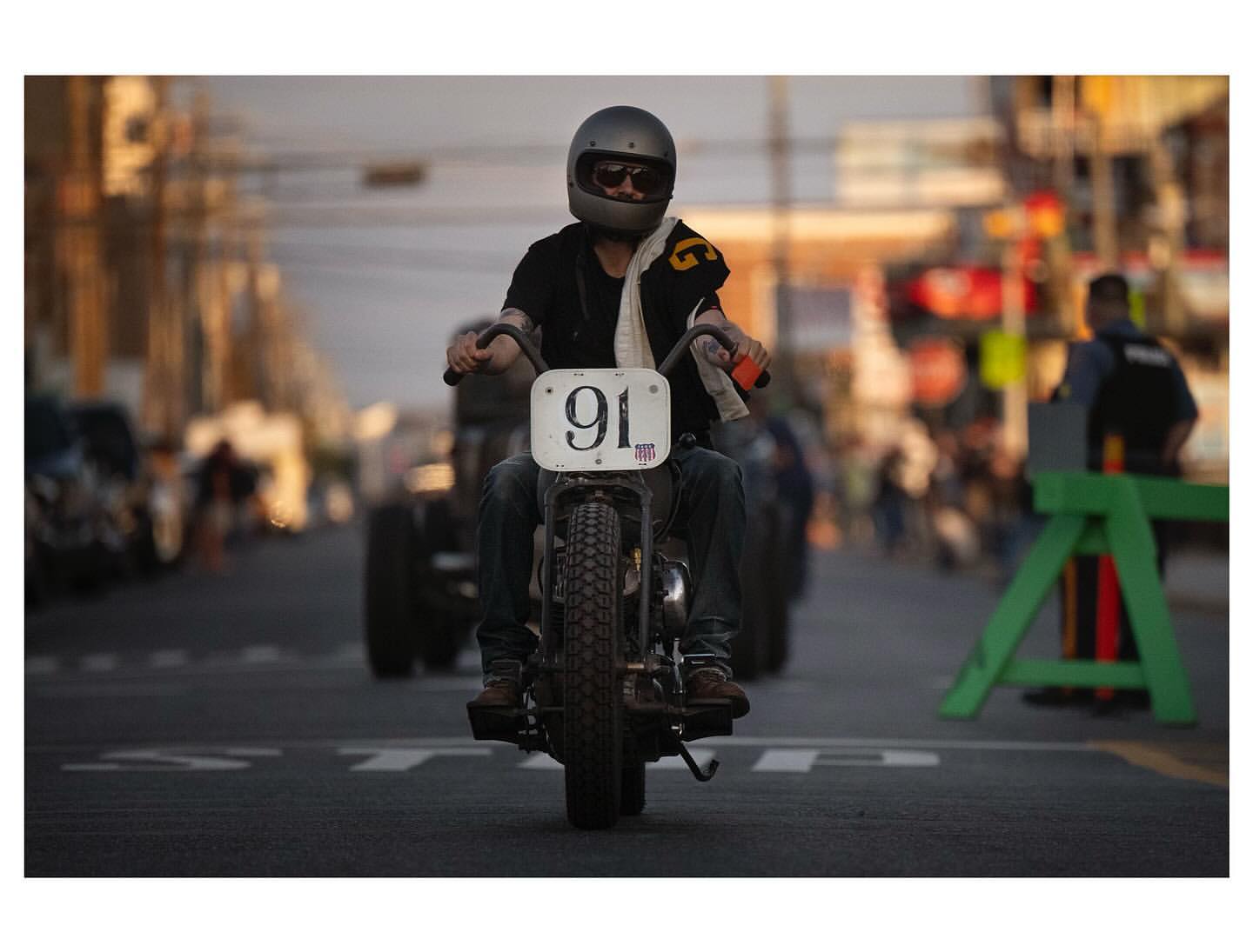 Man riding a vintage motorcycle on a city street, number 91 displayed on the front. People in the background, urban scene.