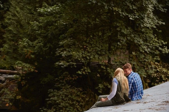 A couple sitting on a rooftop with a forest in the background, the woman has long blonde hair and the man is wearing a blue plaid shirt.