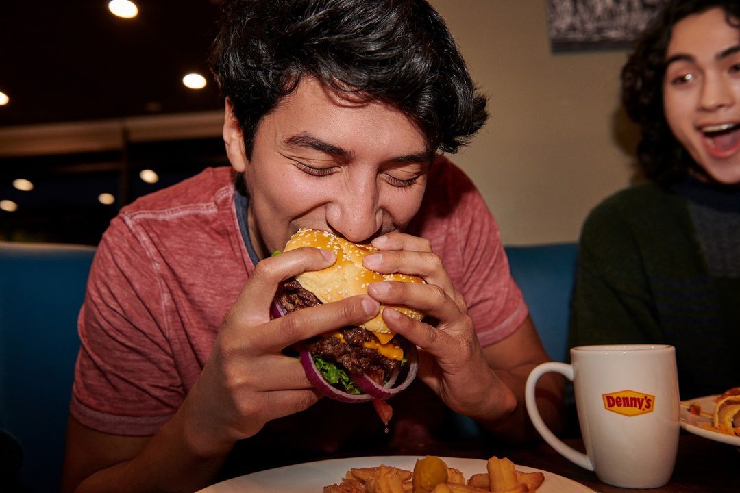 Young man enjoying a burger indoors at a restaurant while another person looks on with excitement, featuring a Denny's mug and fries.