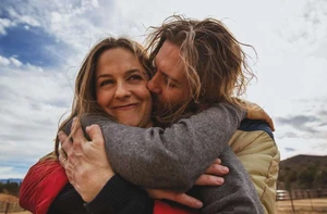 Close-up portrait of a man kissing a smiling woman on the cheek outdoors against a pale sky with soft natural light.
