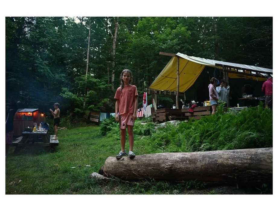 Young girl standing on a fallen log in a forest campsite with tents and people in the background