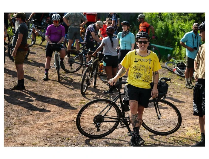 Cyclist in a yellow graphic t-shirt leaning against a black bicycle on a dirt path with a group of cyclists in the background.