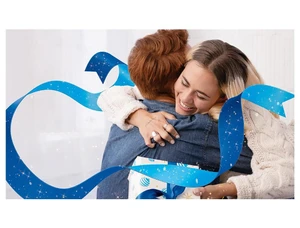 Two people hugging indoors while holding a wrapped gift box, framed by a sweeping digital blue ribbon overlay with sparkling particle effects.
