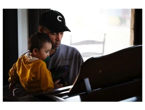 Man wearing black baseball cap holding toddler in yellow top while seated near piano in New York
