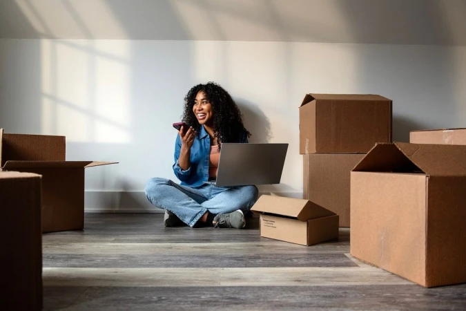 Smiling Black woman sitting cross-legged on a wood floor surrounded by cardboard moving boxes while using a laptop and phone