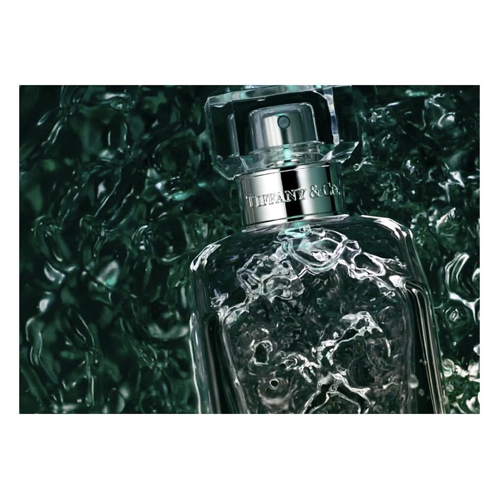 A close-up studio shot showcases a clear Tiffany & Co. perfume bottle against a textured dark green backdrop, emphasizing its luxurious design.