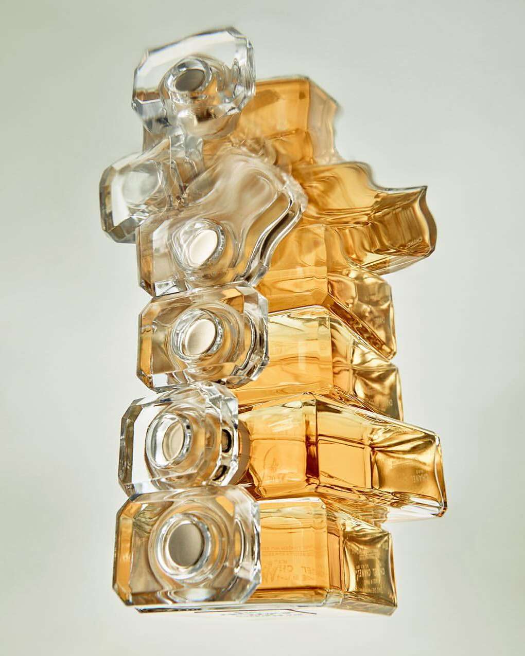 Abstract view of stacked perfume bottles in clear and amber glass creating a geometric sculpture against a plain, light-colored background.