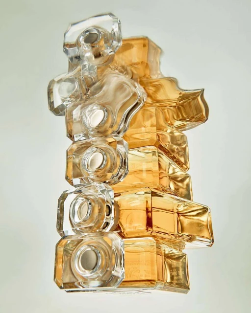 Abstract view of stacked perfume bottles in clear and amber glass creating a geometric sculpture against a plain, light-colored background.