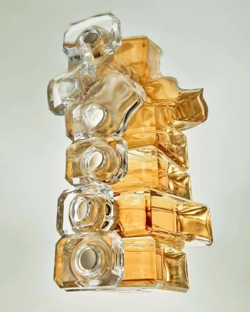 Abstract view of stacked perfume bottles in clear and amber glass creating a geometric sculpture against a plain, light-colored background.