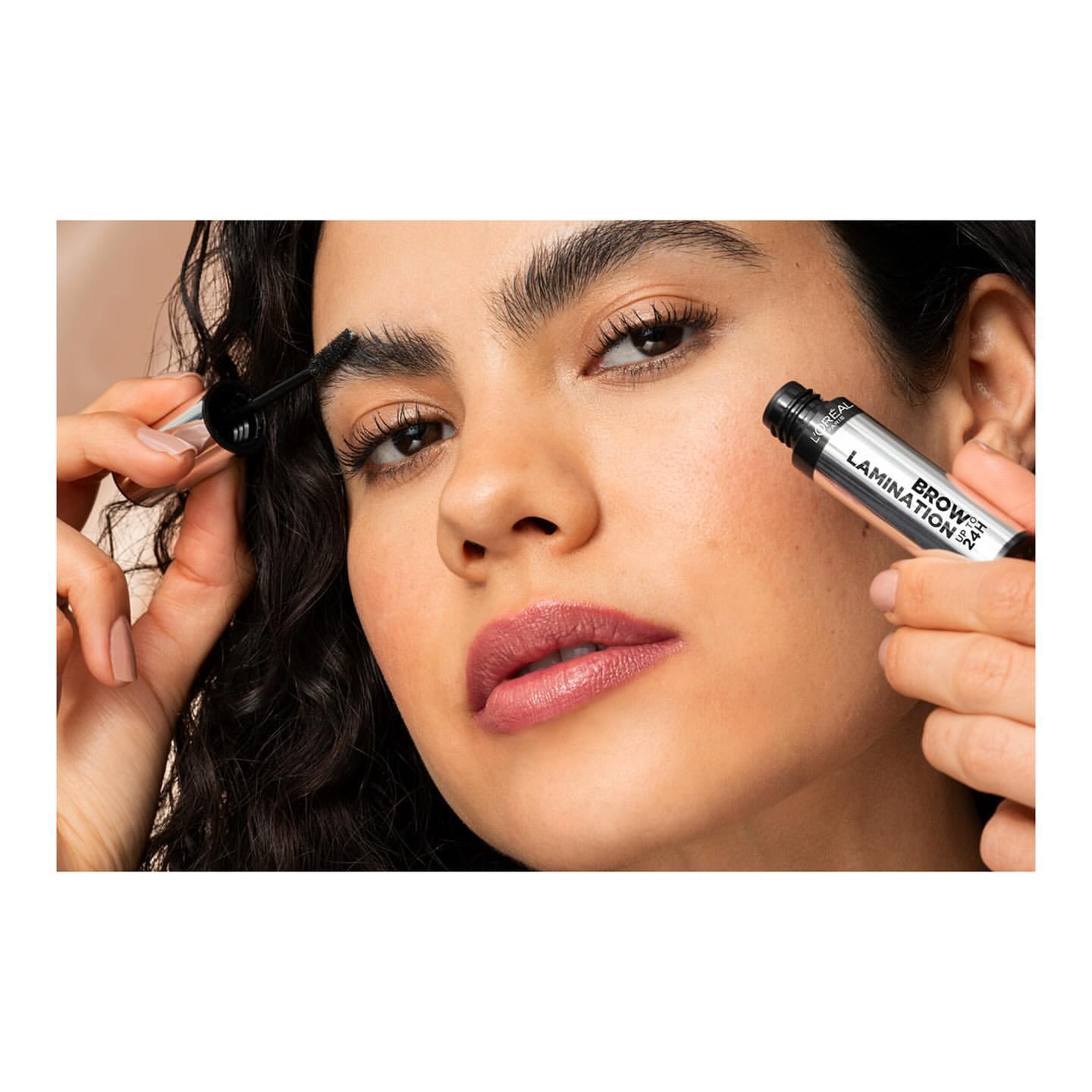 Close-up shot of a woman applying brow gel to her eyebrows. She is holding the silver tube and a wand with a brush on the end.