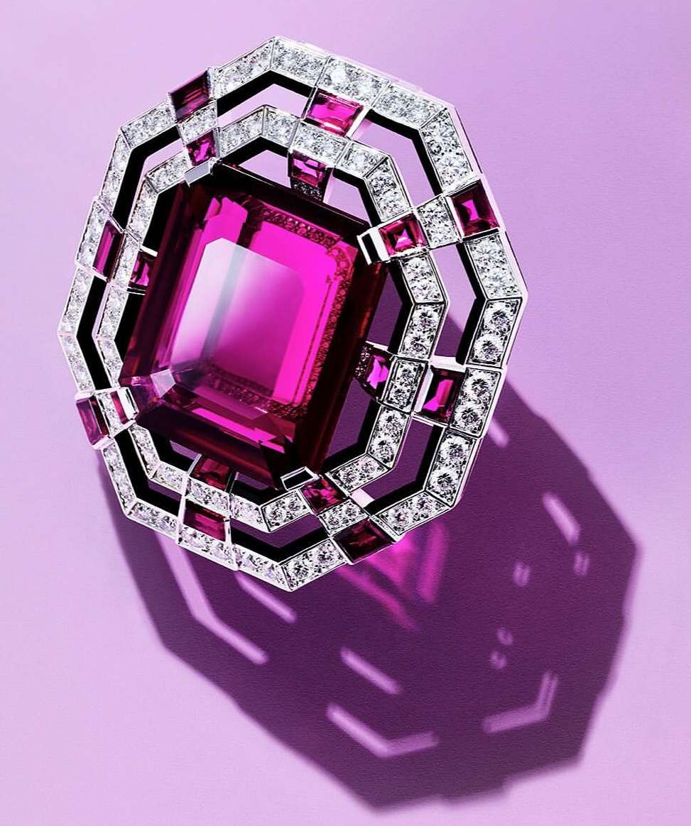 Close-up studio shot of an ornate ring or brooch featuring a large, emerald-cut pink gemstone surrounded by diamonds on a lavender background.