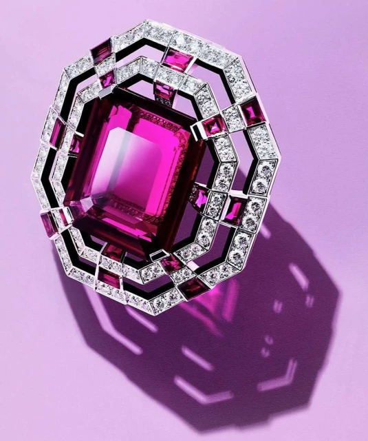 Close-up studio shot of an ornate ring or brooch featuring a large, emerald-cut pink gemstone surrounded by diamonds on a lavender background.
