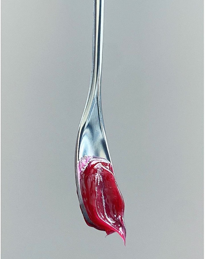 Macro shot of a silver spoon holding thick, glossy, dark red cosmetic cream on a neutral gray background, showcasing textures and color.