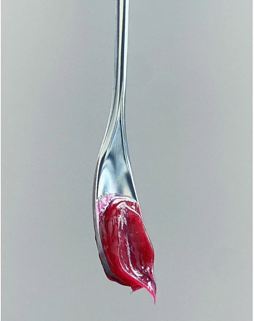 Macro shot of a silver spoon holding thick, glossy, dark red cosmetic cream on a neutral gray background, showcasing textures and color.