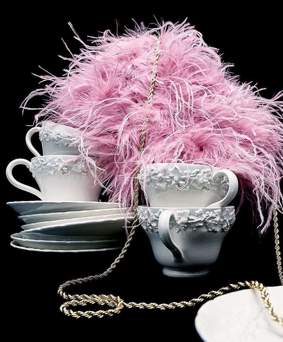 Still life shot of stacked teacups and saucers arranged with pink feathered decorations and draped with gold chain against a black backdrop.