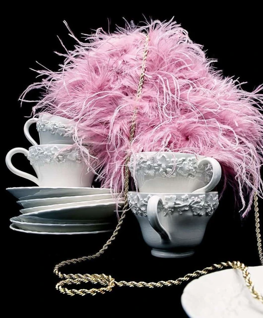 Still life shot of stacked teacups and saucers arranged with pink feathered decorations and draped with gold chain against a black backdrop.