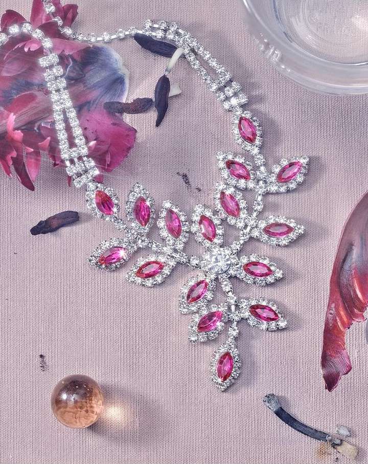 Leaf-shaped rhinestone necklace with pink gems on a textured surface with faint abstract floral patterns and a glass marble.