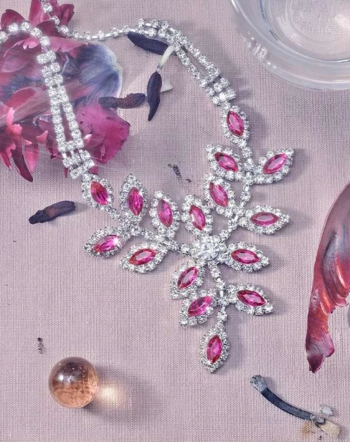 Leaf-shaped rhinestone necklace with pink gems on a textured surface with faint abstract floral patterns and a glass marble.