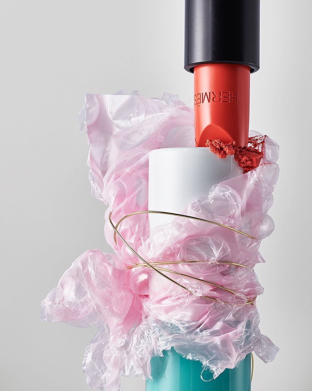 Still life shot of coral lipstick, accessorized with gold wire and pink plastic on top of a cylindrical display on a white background.