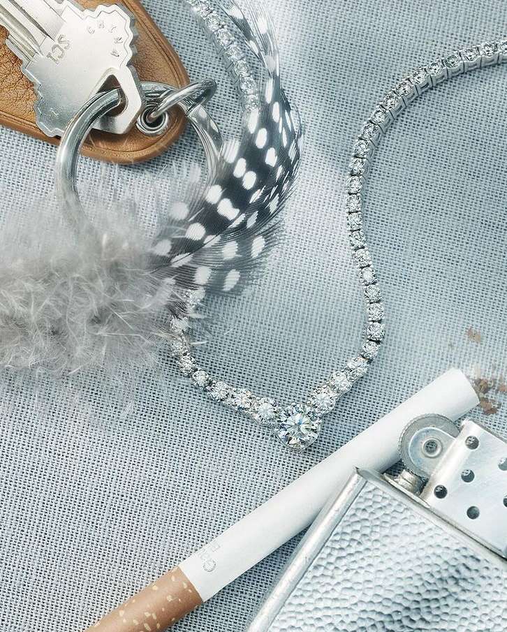 Still life featuring a diamond necklace, cigarette, Zippo lighter, key, and feather on a textured surface, creating a moody, vintage aesthetic.