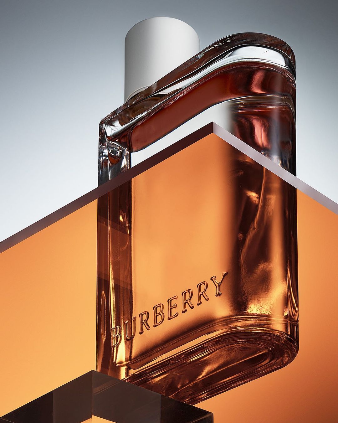A Burberry perfume bottle is staged on a reflective surface, showcasing amber liquid, clear glass, and a cylindrical white cap.